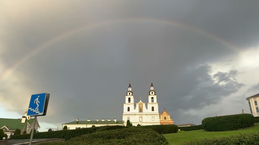 Full rainbow arching over white church with two towers under dark stormy sky, green hedges, blue pedestrian sign, dramatic weather, nature, architecture.