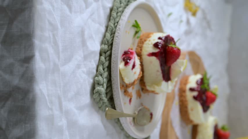 Delicious mini strawberry cheesecakes topped with sweet red berry jam mint and fresh strawberry slices served on a elegant white ceramic plate alongside a small spoon over white linen tablecloth
