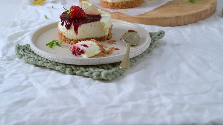 Mini cheesecake topped with rich red berry jam fresh strawberry and lemon slice served on ceramic plate with spoon over rustic white linen background