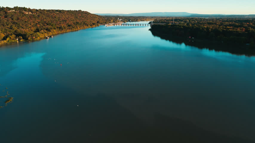 Railroad bridge curves across blue river among autumn trees near Ozark, Arkansas, aerial tracking left with sunset golden glow