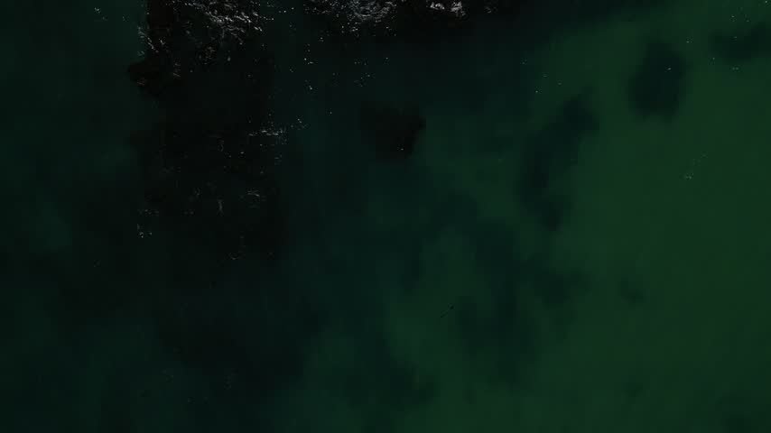 An aerial view of a small island. Flying over one side of the island with the sea breaking against the rocky shoreline.