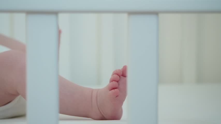 Baby moves legs behind crib bars, creating visual image of protected home space where everyday care, safe rest, and natural physical activity come together during early infancy indoors.