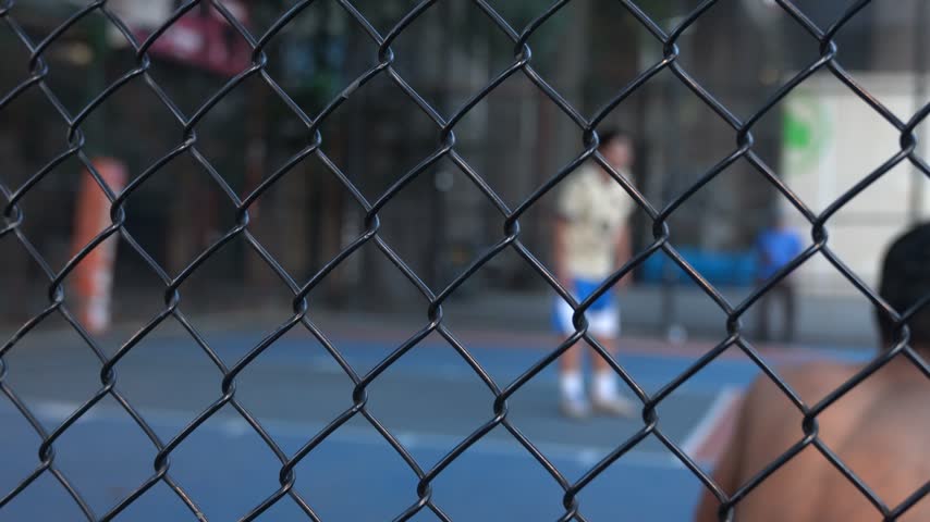Stock video footage of people playing a basketball game outdoors, filmed through a chain-link fence on an urban sports court.
