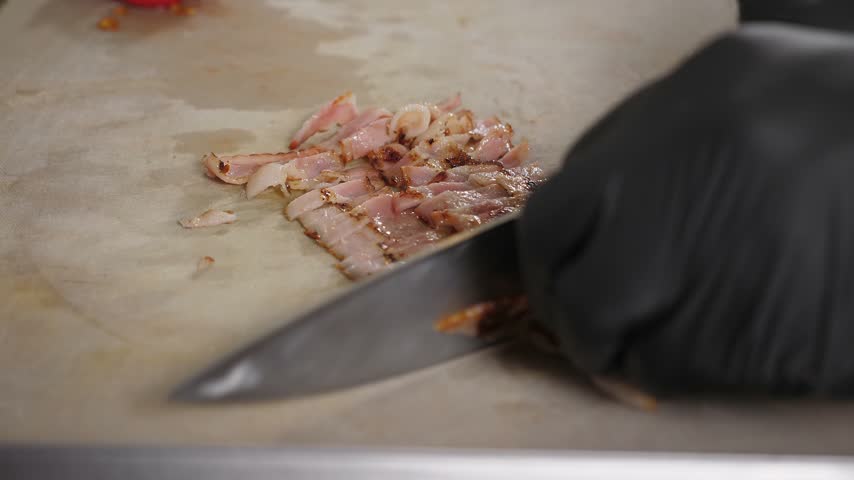 A close-up of a chef wearing gloves, slicing fried bacon with a knife on a cutting board. The process of cooking fast food in the kitchen of the restaurant.