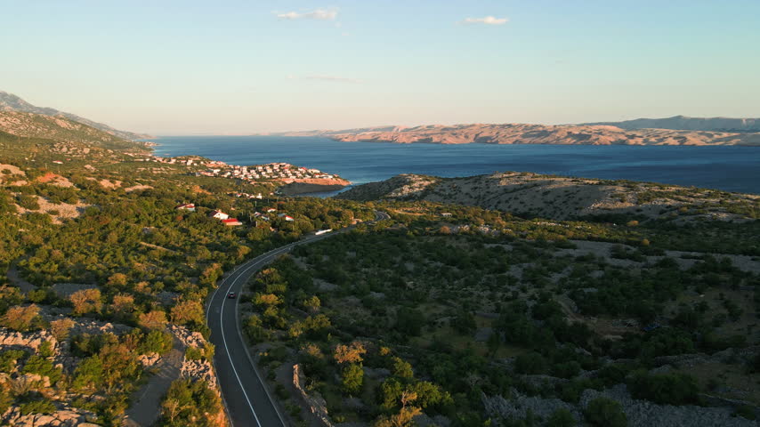 Car driving along a coastal road near the sea, surrounded by rocky green hills and a small village. Aerial view of scenic travel through a coastal landscape.