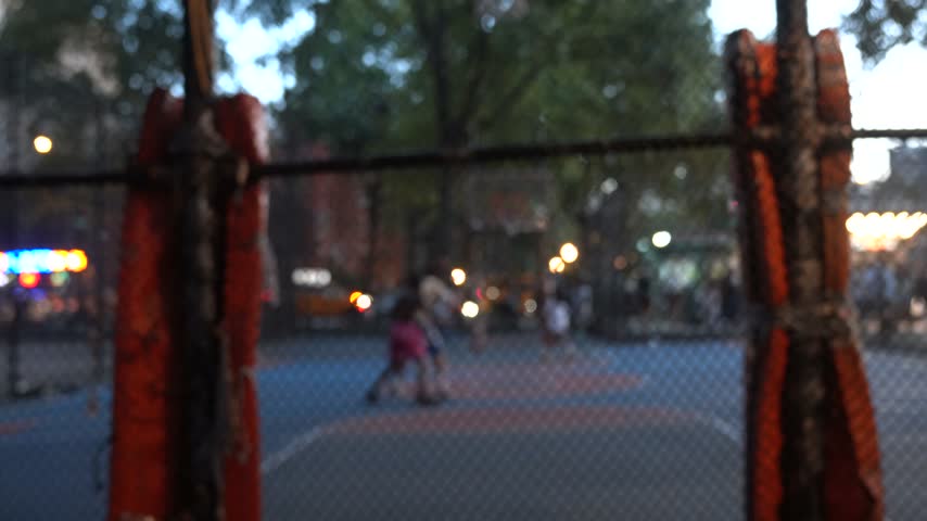 Stock video footage of people playing a basketball game outdoors, filmed through a chain-link fence on an urban sports court.

