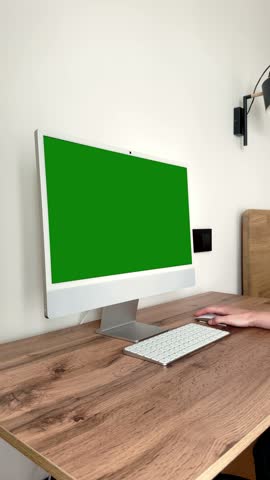 Chroma key screen computer display resting on wooden table. Freelance employee hand holding mouse controller near office hardware accessories. Ambient room lighting fills cozy home office background