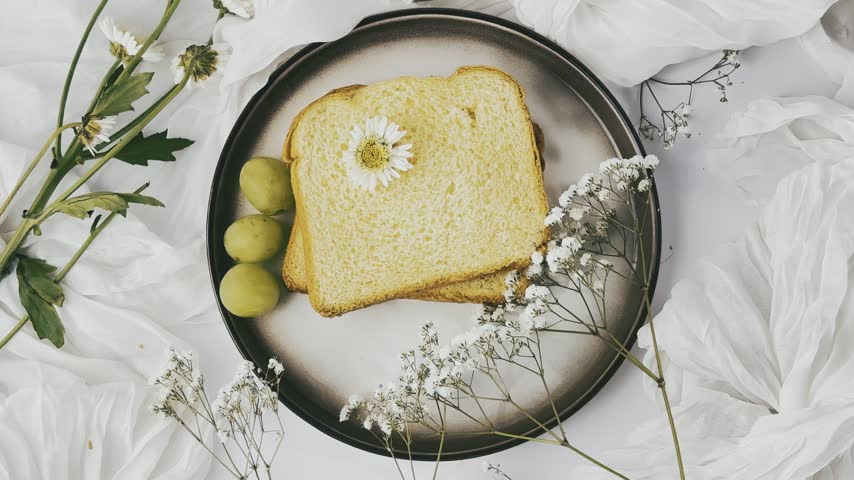 Toasted bread, three green grapes, delicate blossoms, and a small white flower are arranged on a round plate surrounded by softly folded pale cloth.