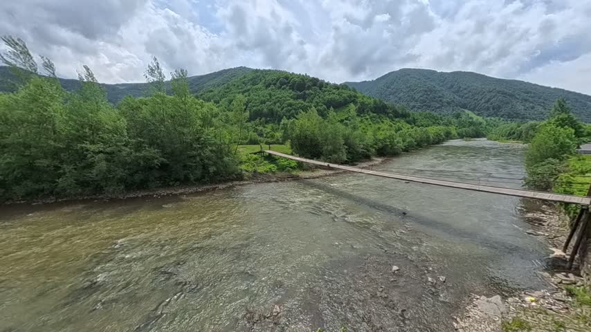 First-person POV perspective looking across a narrow wooden suspension bridge crossing a wide, flowing mountain river towards lush green forested hills under a cloudy sky. A wooden suspension bridge over a fast mountain river.