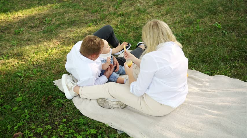 Cute baby eating during family picnic in summer park, toddler sits with dad and eats cookie, while mother sits nearby holding small jar of baby puree. healthy baby snacks and organic food concept.