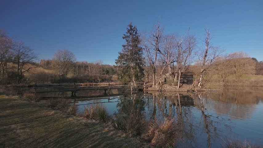 Scenic landscape view of a calm lake with a small island, bare trees, and a wooden bridge leading to a small hut under a clear blue sky