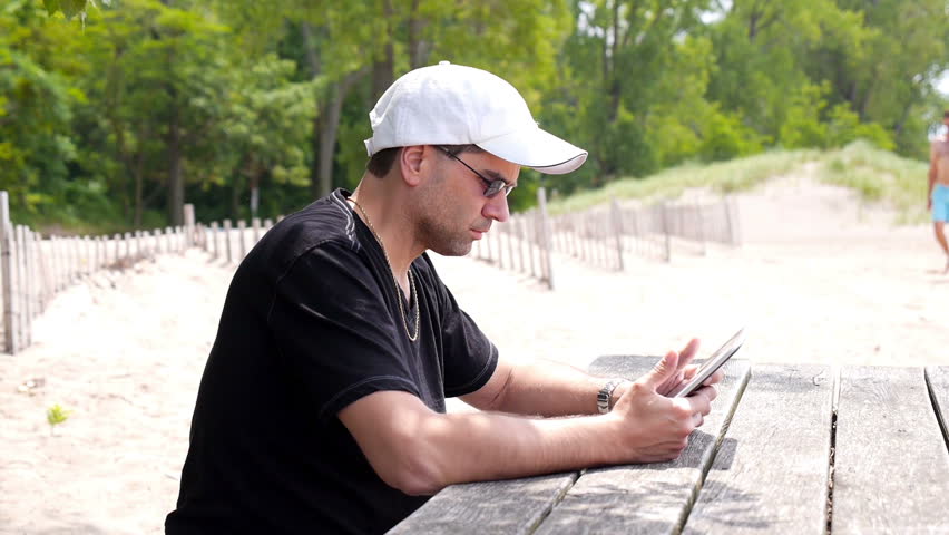 A man at the beach uses his tablet PC at a beachside picnic table.