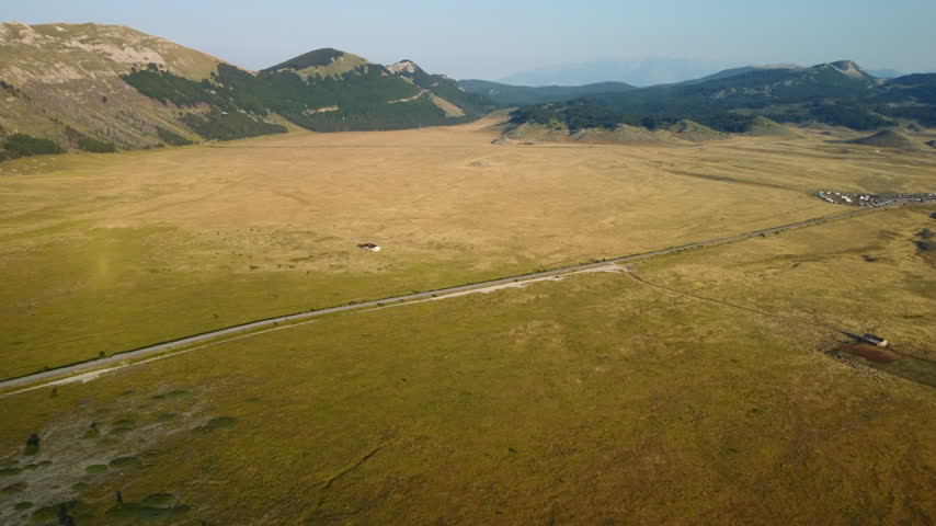Drone mountain road yellow meadow, forward aerial flight. Vast flat treeless barren grassland with isolated house and wooded peaks background.