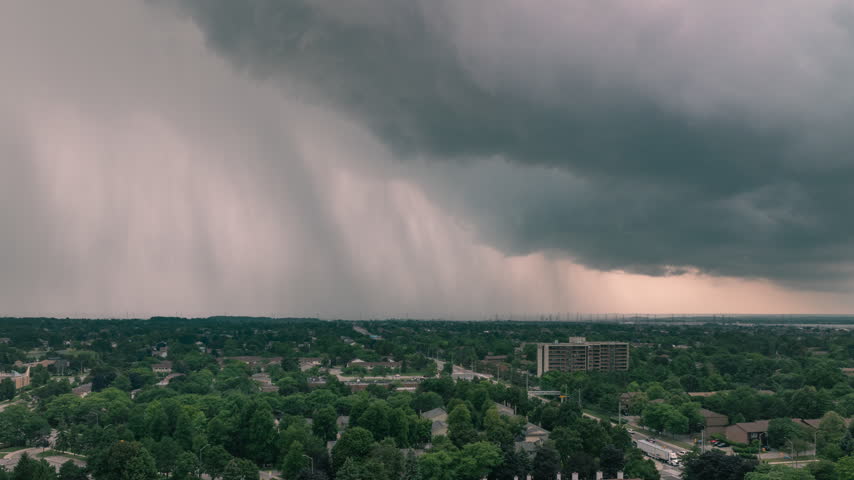 Heavy rain falls from dark storm clouds over a suburban residential neighborhood in Mississauga, Ontario. The fast-moving storm creates a visible downburst over the green landscape.