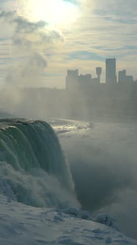 Vertical winter view of American Falls plunging through dense mist, with turquoise water, snow-covered frozen rocks and the Canadian skyline at Niagara Waterfalls, Canada