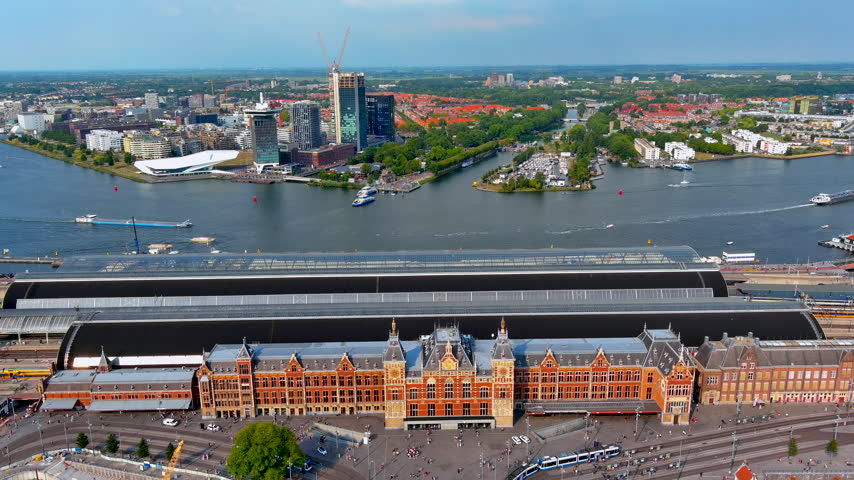Aerial drone view of Amsterdam Centraal railway station with trams and travelers on Stationsplein, platform roofs, ferries on the IJ river and the Amsterdam Noord skyline, Netherlands.