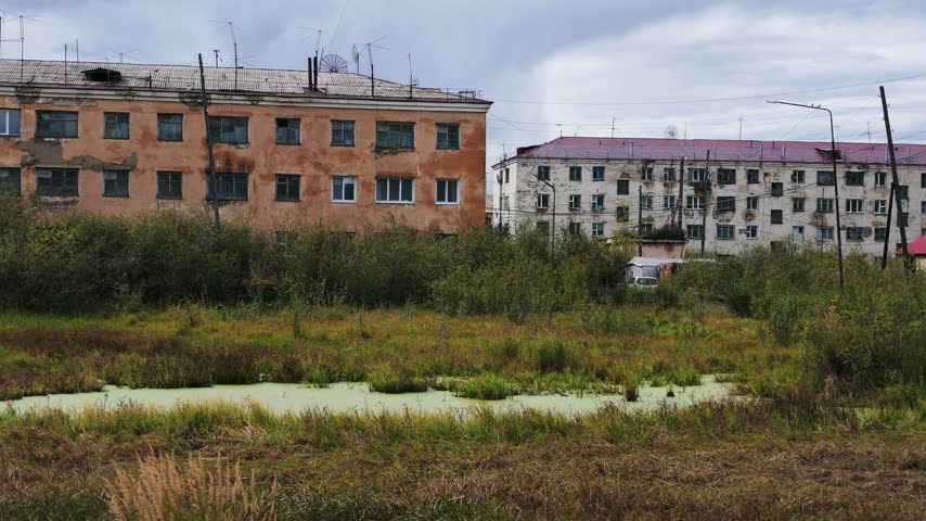Two weathered apartment buildings stand behind overgrown vegetation and a stagnant, algae-covered pond under a cloudy sky, with no visible movement or change throughout the sequence.