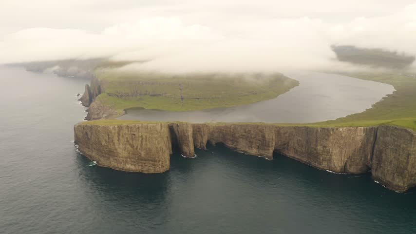 Aerial view of Sorvagsvatn lake perched on steep cliffs above the ocean with low hanging clouds and green grassy hills of Faroe Islands.