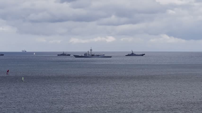 Aerial view of military ships sailing on the horizon in Sopot, Poland with a windsurfer on the water under cloudy skies.