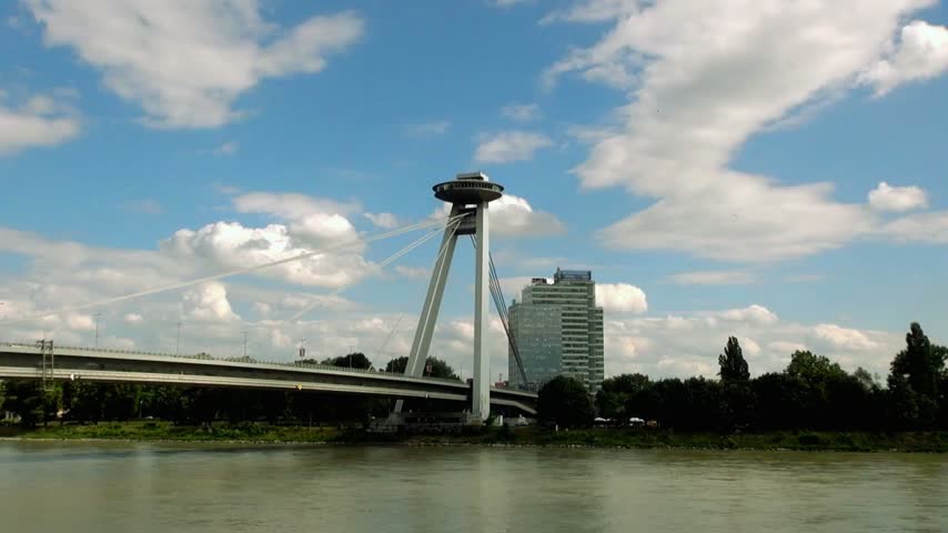 Aerial view of New Bridge in Bratislava, Slovakia, spanning the Danube River on a cloudy day, featuring modern architecture, urban surroundings, river traffic, and a distinctive European cityscape.