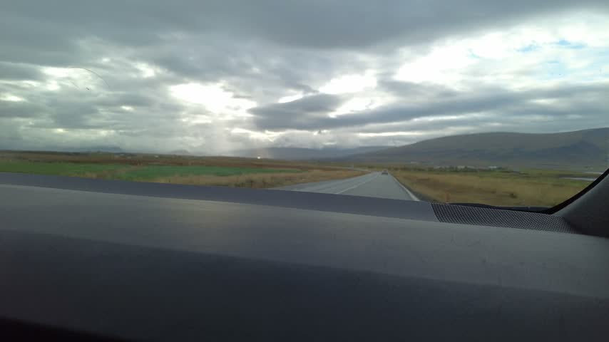 Driving Through Rural Iceland Under Dramatic Cloudy Sky - POV car dashboard view traveling a remote Icelandic highway. Sun rays pierce heavy clouds over distant mountains and open fields.