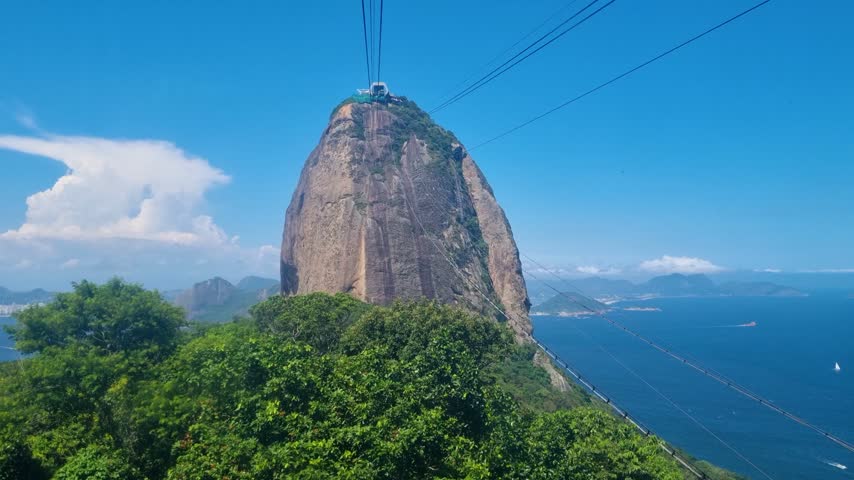 Forward-moving cable car view of Sugarloaf Mountain, Pão de Açúcar, in Rio de Janeiro, Brazil, rising above lush tropical vegetation beneath a clear blue sky at the iconic attraction