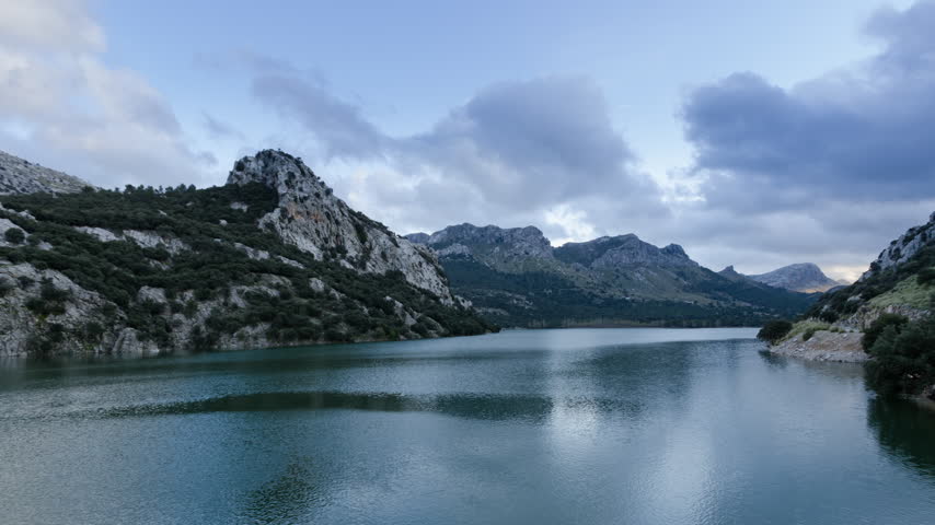 Static view of Gorg Blau reservoir surrounded by rocky mountains in Mallorca, Spain, beneath a cloudy sky.