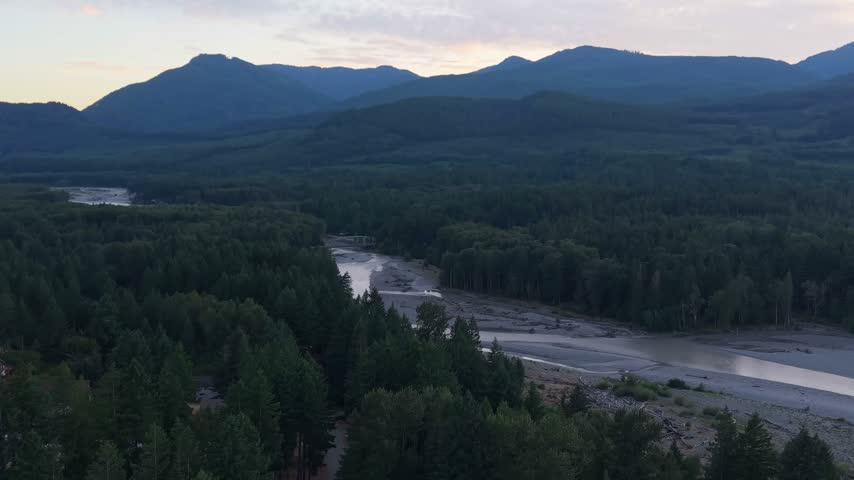 Aerial view of the Cowlitz River winding through a forested landscape near Packwood, Washington, with Mount Rainier and surrounding mountain ridges visible in the distance during evening light.
