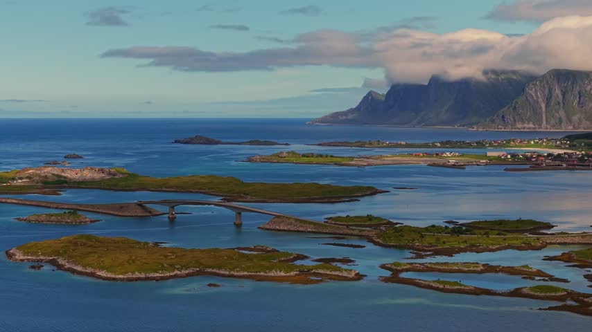 Drone view centered on Henningsvær Bridge, linking the island fishing village across clear coastal waters, with rocky islets and roads in Norway’s Lofoten archipelago