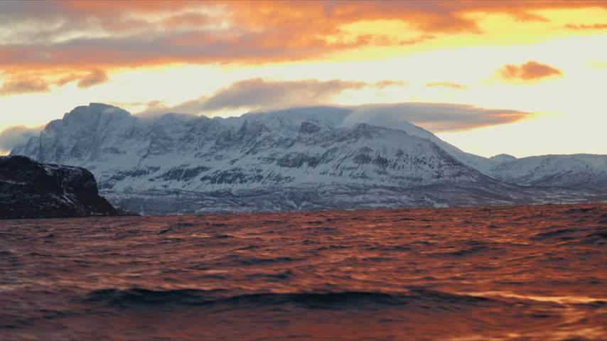Snow covered Arctic mountains rise beyond orange sunset light on the cold Norway sea