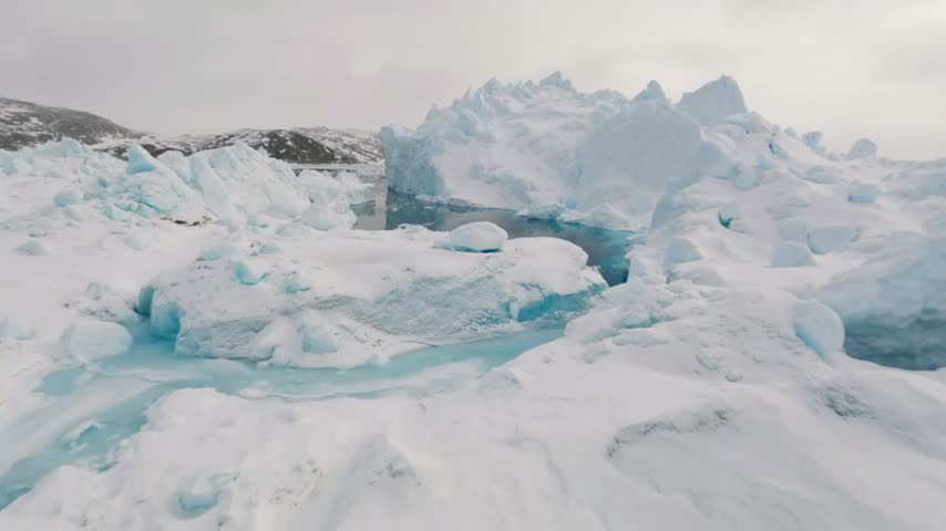 Fast and dynamic aerial drone flight navigating low over calm, dark blue water through massive icebergs and floating ice chunks in a frozen polar landscape