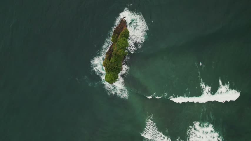 Aerial top-down shot of a small, lush green rocky islet surrounded by dark ocean waters, capturing white foam waves crashing against the islet cliffs and rolling past surfers waiting in the sea.