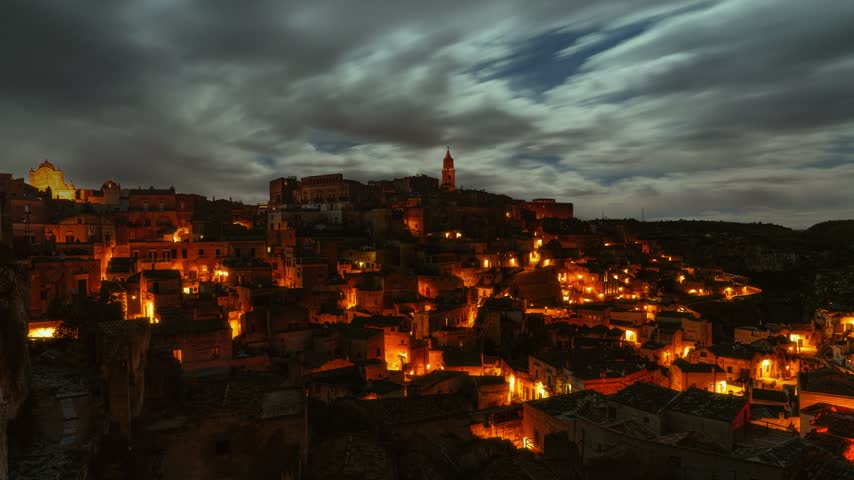 Night time lapse of moving clouds and stars over illuminated Sassi. Ancient old town of Matera in Italy.