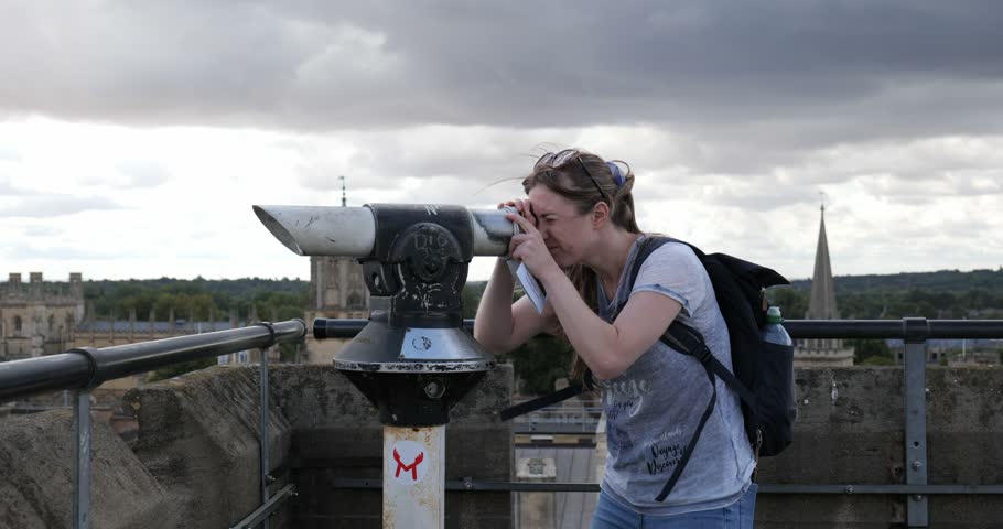 Oxford, Oxfordshire, United Kingdom, August 20, 2026. Side view of a young tourist woman looking through a telescope on top of Carfax Tower, observing the historic city skyline under a cloudy sky.