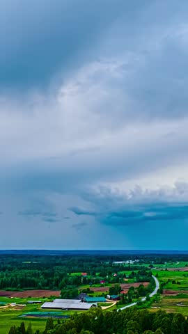 Aerial vertical timelapse captures fast moving storm clouds over rural farm buildings and green fields at sunset in Latvia