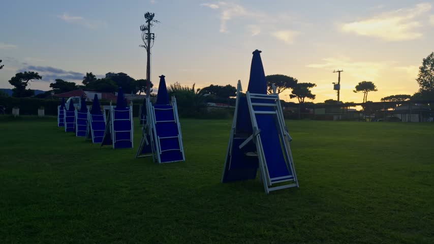 Row of empty, folded blue beach chairs on a green lawn during a beautiful sunset. End of the summer season concept with no people