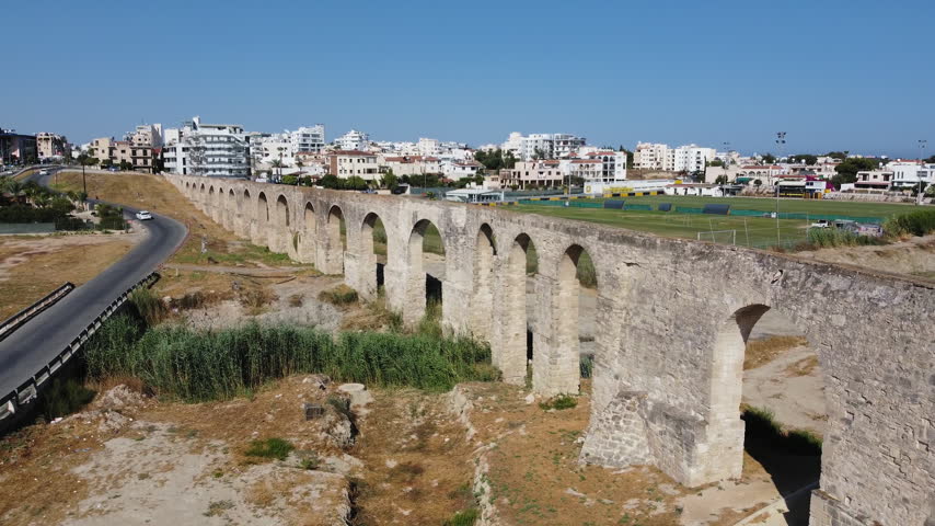 Stone arches of an old aqueduct curve alongside a road with modern buildings in the background under a clear blue sky. Ancient arches beside a roadway.