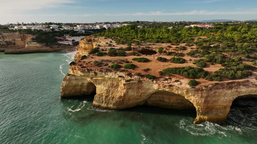 Drone view of cliffs above Benagil Cave in Algarve, Portugal, with limestone sea caves, turquoise Atlantic water, waves, and rugged coast. Orbit parallax shot.