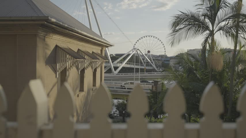A heritage cottage sits behind a white picket fence with palm trees and a distant Ferris wheel in Brisbane.