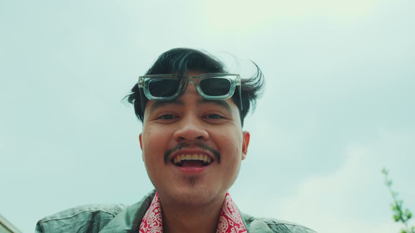 A smiling young Asian man in casual streetwear holding the camera, looking directly into the lens and talking enthusiastically.