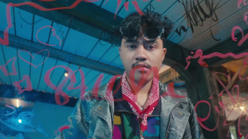 A trendy young Asian man with a mustache, sunglasses, and red bandana looks intensely at the camera through a glass pane covered in red marker writing.