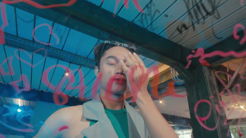 Close-up portrait of a stylish young man posing confidently with his hand in his hair, looking intensely through a glass window covered in red marker writing.
