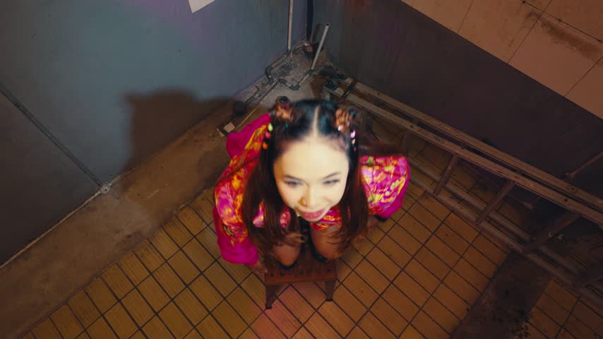 High angle portrait of a cute asian woman with twin buns in a colourful jacket looking up at the camera playfully under vibrant pink neon lighting.
