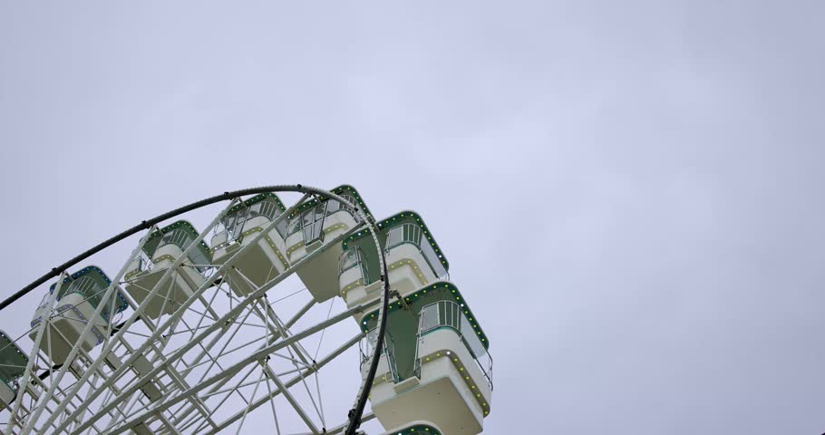 Slow motion footage in Llandudno North Wales showing a large illuminated Ferris wheel from below with passenger gondolas metal framework and decorative lights against a cloudy sky at the seaside