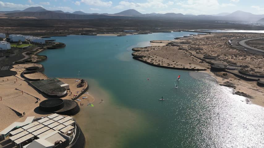 Calm blue lagoon curves between sandy shores with volcanic mountains beyond, aerial low descend flyover of paddleboarders on glistening water