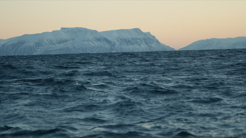Snow covered Arctic mountains rise beyond the choppy Norway sea under a pale twilight sky