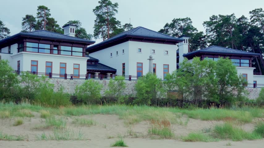 Medium view of elegant contemporary seaside mansion with white walls and dark roofs behind sand dune vegetation in Jurmala resort town on Baltic coast, Latvia.