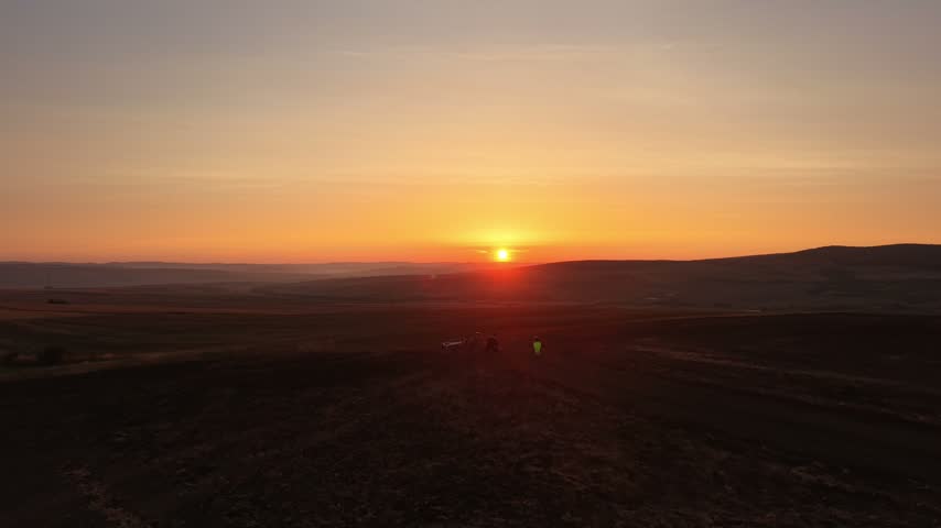 Forward drone flight passing over two cyclists stopping to watch the sunrise before continuing toward the glowing morning sky.