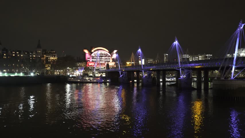 Static tripod shot of the illuminated Golden Jubilee Bridge over the River Thames in central London at night. Colorful city lights reflect across the water, with the distinctive Charing Cross area arc
