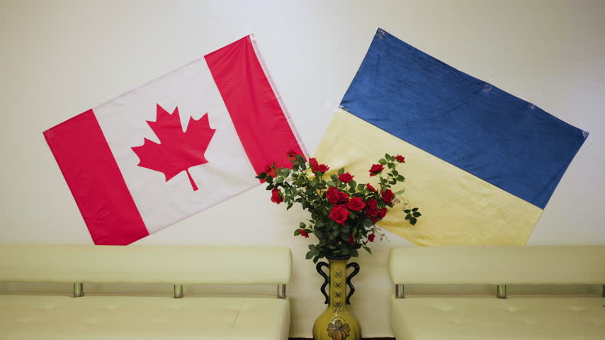 Two flags are displayed on a wall. The Canadian flag is on the left, and the Ukrainian flag is on the right. A vase with red roses sits on a table below the flags.